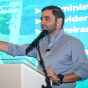 Homem de camisa azul claro fala ao microfone em um palco, gesticulando com a mão. Atrás dele, há uma tela grande com texto em português e a imagem de uma estrada. Parece ser uma apresentação ou conferência.