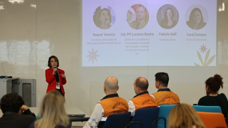 Imagem de uma apresentação institucional em ambiente de conferência. Uma pessoa está em pé, falando ao microfone, vestindo blazer vermelho. Ao fundo, há um slide projetado na parede com o título “ABERTURA INSTITUCIONAL”, que exibe quatro fotos com nomes e cargos:

Raquel Teixeira – Secretária de Estado da Educação do RS
Cel. PM Luciano Boeira – Chefe da Casa Militar e Coordenador Estadual de Proteção e Defesa Civil do RS
Fabiola Galli – Instituto Alana
Carol Campos – Vozes da Educação

Na plateia, algumas pessoas estão sentadas, incluindo participantes com coletes laranja com a inscrição “DEFESA CIVIL” nas costas. O ambiente sugere um evento formal voltado à educação e gestão de riscos climáticos.