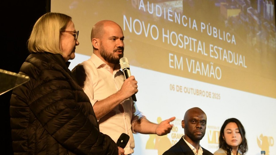 Quatro pessoas estão participando de um evento público. No centro da imagem, um homem de camisa social branca segura um microfone e está falando. Ao lado esquerdo dele, uma mulher mais velha, com cabelos loiros e lisos, usa óculos e uma jaqueta preta. À direita, estão sentados um homem negro, careca, usando terno preto e camisa branca, e uma mulher jovem de cabelos lisos e escuros, com uma jaqueta clara.
Ao fundo, há uma tela de projeção com os dizeres:
"Audiência Pública - Novo Hospital Estadual em Viamão", com a data 06 de outubro de 2025.
Também aparecem os logotipos do Governo do Estado do Rio Grande do Sul, da Prefeitura de Viamão e do SUS.
O clima do evento é formal, com foco em uma discussão pública sobre a construção de um novo hospital estadual na cidade de Viamão, no Rio Grande do Sul.
