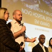 Quatro pessoas estão participando de um evento público. No centro da imagem, um homem de camisa social branca segura um microfone e está falando. Ao lado esquerdo dele, uma mulher mais velha, com cabelos loiros e lisos, usa óculos e uma jaqueta preta. À direita, estão sentados um homem negro, careca, usando terno preto e camisa branca, e uma mulher jovem de cabelos lisos e escuros, com uma jaqueta clara.
Ao fundo, há uma tela de projeção com os dizeres:
"Audiência Pública - Novo Hospital Estadual em Viamão", com a data 06 de outubro de 2025.
Também aparecem os logotipos do Governo do Estado do Rio Grande do Sul, da Prefeitura de Viamão e do SUS.
O clima do evento é formal, com foco em uma discussão pública sobre a construção de um novo hospital estadual na cidade de Viamão, no Rio Grande do Sul.