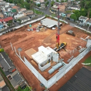 A imagem mostra uma vista aérea de um canteiro de obras em estágio avançado, localizado em uma área urbana cercada por residências. O terreno é amplo, com solo avermelhado, e já conta com muros de concreto. No interior, há estruturas em construção, guindastes e veículos de apoio, além de áreas destinadas a materiais e equipamentos.