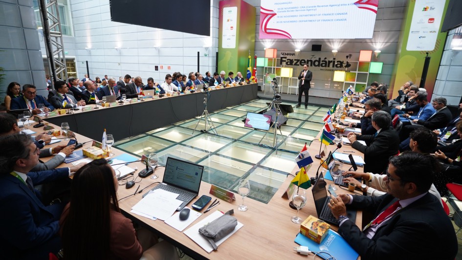 Foto mostra palestrante em pé, em frente a um painel que diz "Semana Fazendária", numa sala com uma grande mesa em formato de U, na qual estão dispostos dezenas de representantes dos estados brasileiros. Na mesa na frente de cada representante há uma pequena bandeira de seus Estados. 