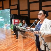A imagem destaca uma mulher, titular da Sema, que fala ao microfone em um púlpito transparente. Ao fundo, aparecem outras duas mulheres sentadas. Também há um telão com o título do evento: 1º Encontro Estadual pelo Dia dos Animais.
