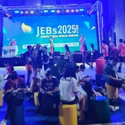 Foto mostra ambiente interno iluminado por luzes coloridas, com jovens sentados em pufes de diferentes cores diante de um palco. No telão ao fundo, aparece o texto “JEBs 2025 Uberlândia - Minas Gerais”. Pessoas circulam e conversam no local.