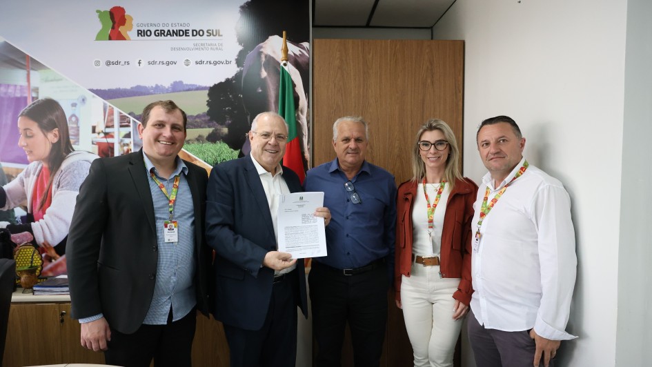 A imagem mostra um grupo de cinco pessoas em um ambiente interno, posando para uma foto institucional. No centro, um homem segura um documento aberto. Ao fundo, há um painel com a identidade visual do Governo do Estado do Rio Grande do Sul.