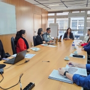 A imagem mostra uma reunião em uma sala de conferências. Nove pessoas estão sentadas ao redor de uma mesa retangular de madeira clara. Sobre a mesa há laptops, papéis, canetas e xícaras de café. A sala possui paredes revestidas de madeira e grandes janelas ao fundo, que permitem a entrada de luz natural. Uma pessoa está falando enquanto as demais escutam. À esquerda, há um quadro branco na parede, e próximo às janelas, um flip chart.