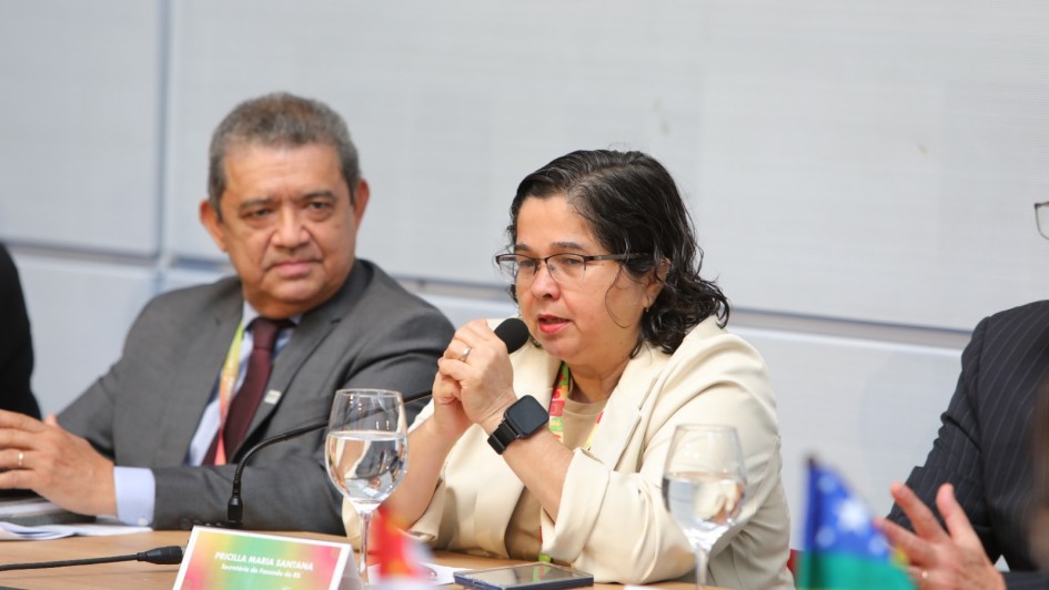 A imagem mostra uma mulher e dois homens sentados à mesa de uma conferência ou reunião formal. A mulher, que está no centro da imagem, fala ao microfone. Ela usa óculos, um blazer claro, e um relógio inteligente preto no pulso esquerdo. Em sua frente há um copo de água, um celular e uma placa de identificação com o nome "Priscila Maria Santana", identificando-a como "Secretária de Fazenda do ES" (Espírito Santo). À sua direita (à esquerda na imagem), está um homem de terno escuro e gravata, observando atentamente. À esquerda dela (direita da imagem), aparece parcialmente outro homem, também de terno. Na mesa estão bandeiras de estados brasileiros, incluindo a de São Paulo e Tocantins. O ambiente é bem iluminado, com um fundo neutro.