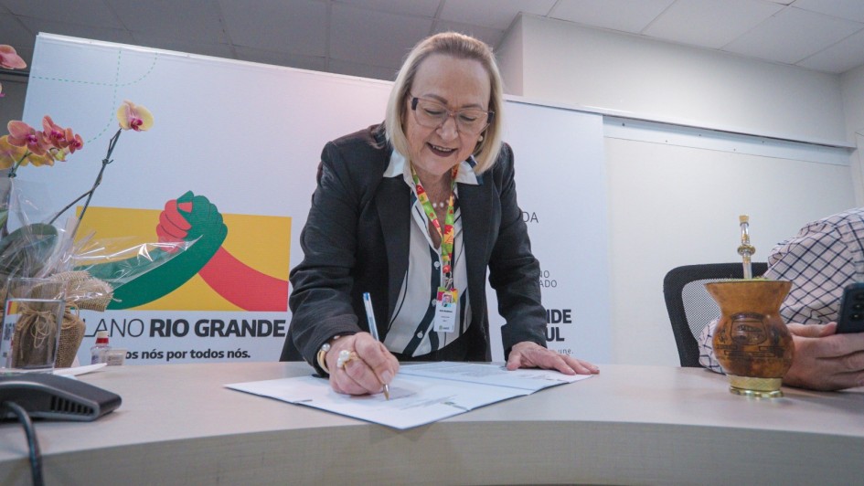 Foto mostra secret&aacute;ria Arita Bergmann assinando documento. Atr&aacute;s dela, h&aacute; um banner do Plano Rio Grande.