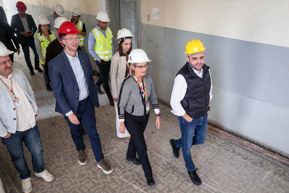 Um grupo de pessoas caminha por um corredor, usando capacetes de segurança e roupas apropriadas para visita a obra. Algumas estão vestidas de forma formal, com ternos, enquanto outras usam trajes mais casuais. O piso é revestido com cerâmica, e as paredes são pintadas em dois tons de cinza — claro na parte superior e escuro na inferior. A cena indica uma inspeção ou visita técnica a um local em reforma ou construção.