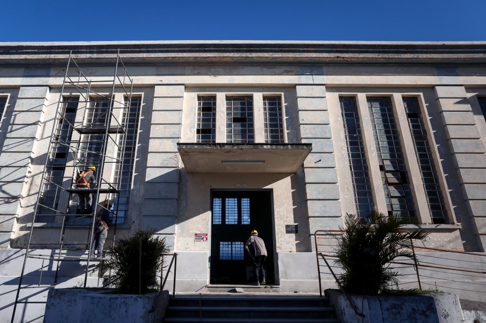 A imagem mostra a fachada de um edifício antigo em reforma. À esquerda, há andaimes onde dois trabalhadores com capacetes realizam reparos. No centro, uma porta preta está aberta e uma pessoa está posicionada em frente a ela. O prédio possui janelas altas e estreitas com grades metálicas, e a fachada apresenta sinais de desgaste, como pintura descascada e manchas. Há duas plantas em vasos grandes nas laterais da escada que leva à entrada principal. O céu está claro e azul, indicando um dia ensolarado.