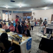 A imagem mostra um ambiente interno, parecido com um salão comunitário, onde está ocorrendo um mutirão de atendimento ao público. Diversas pessoas estão sentadas ou em pé, aguardando ou sendo atendidas em mesas com computadores. O espaço está organizado com mesas distribuídas em fileiras, e há placas com nomes de serviços como "Registro de imóveis", "Projeto Terra" e "Justiça". Algumas pessoas estão com pastas ou documentos nas mãos. Os atendentes estão concentrados nos computadores, realizando cadastros ou tirando dúvidas. O público é composto majoritariamente por adultos e idosos. O ambiente é simples, com piso de cerâmica e teto de madeira, e há ventiladores fixados nas paredes. Ao fundo, há mais pessoas e uma porta aberta por onde entra luz natural. A atmosfera é de organização e prestação de serviços comunitários.