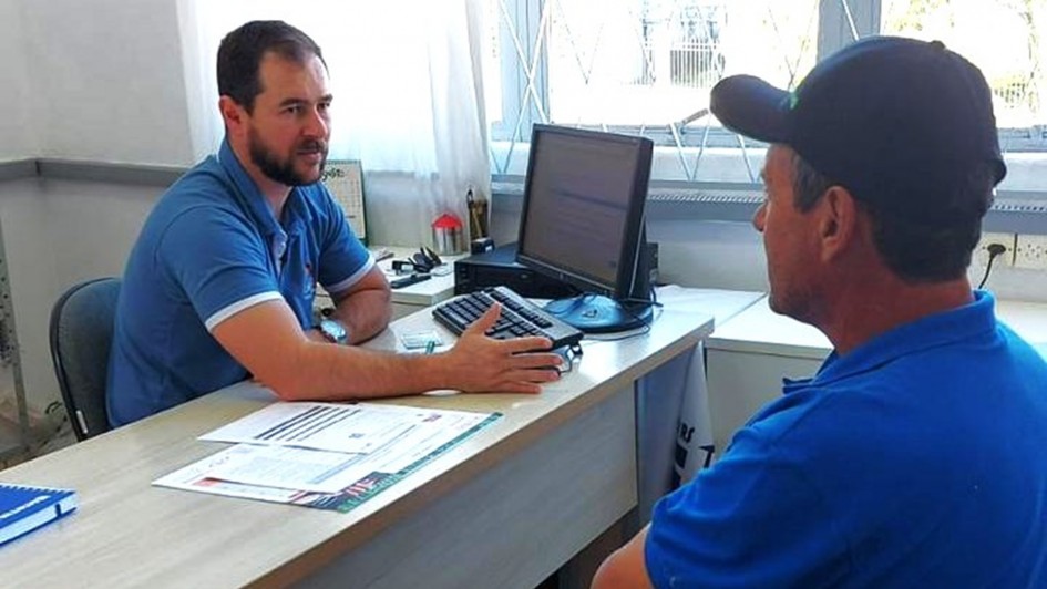 Duas pessoas estão sentadas em lados opostos de uma mesa em um ambiente que se assemelha a um escritório. A pessoa à esquerda, vestindo uma camisa azul, gesticula com a mão direita enquanto conversa com a outra. Sobre a mesa, há diversos papéis, um teclado, um mouse e um monitor de computador. A pessoa à direita também usa camisa azul e boné preto. Ao fundo, vê-se uma grande janela com grades e cortinas brancas.