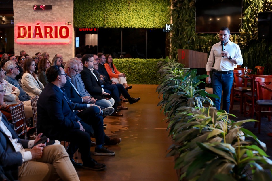 A imagem mostra o vice-governador diante da plateia. As pessoas estão sentadas em cadeiras transparentes, assistindo a apresentação.