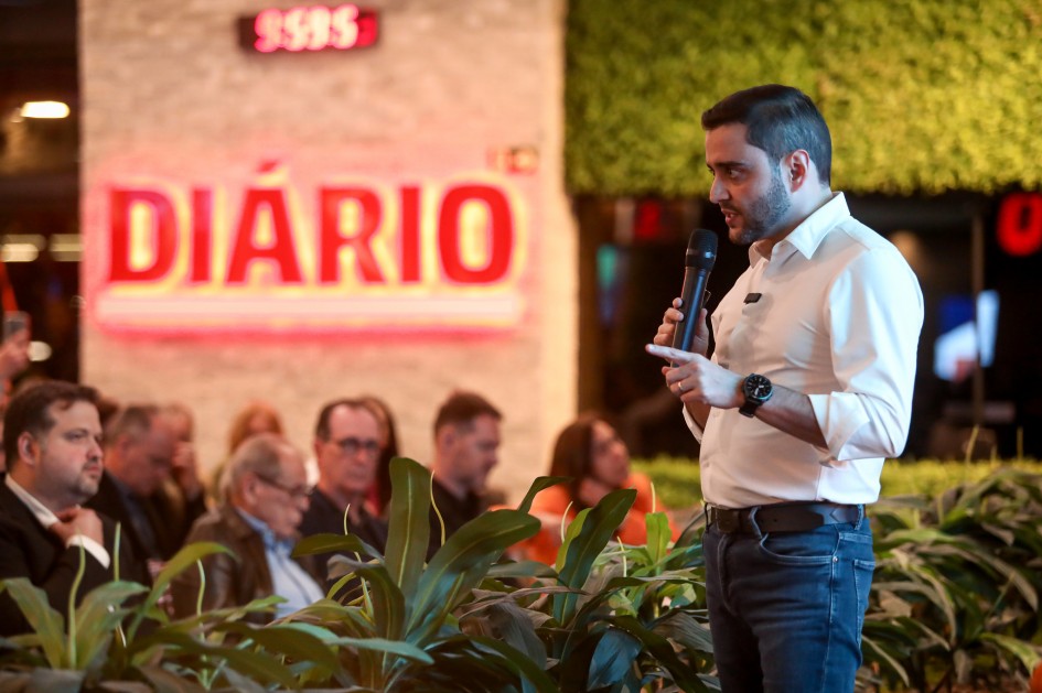 A imagem mostra um ambiente interno com decoração moderna, onde ocorre um evento com público sentado em cadeiras organizadas em fileiras. Em primeiro plano, o vice-governador está em pé segurando um microfone.