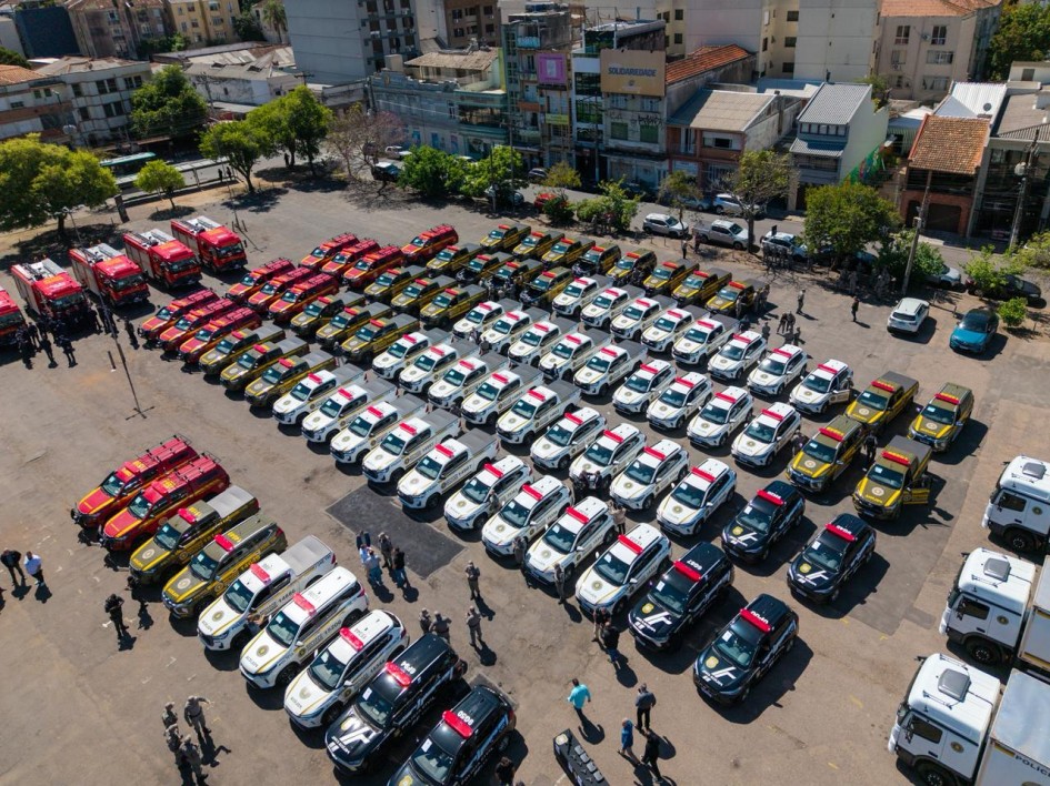 A imagem mostra um grande pátio com dezenas de viaturas novas alinhadas, vistas de cima. Há veículos brancos, pretos, amarelos e vermelhos, incluindo caminhonetes e caminhões de bombeiros. Algumas pessoas estão em pé próximas aos carros, aparentemente em um evento oficial de entrega de veículos para forças de segurança pública. Ao fundo, há prédios e árvores de uma área urbana.