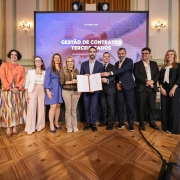 Um grupo de 12 pessoas, formado por homens e mulheres, posa sorridente para uma foto em um ambiente formal, com decoração elegante e piso de madeira trabalhada. Todos estão vestidos com roupas sociais, adequadas a um evento corporativo ou institucional. Ao centro, duas pessoas seguram um documento oficial aberto, emoldurado com capa vermelha, como um certificado ou contrato.
Ao fundo, há uma tela grande com o texto:
"OUTUBRO | 2025
GESTÃO DE CONTRATOS TERCEIRIZADOS
Novas regras para a gestão contratual", indicando que o evento se relaciona com mudanças ou diretrizes em contratos terceirizados. O ambiente parece ser um salão de eventos governamental ou institucional, com cortinas douradas, colunas e lustres de parede.