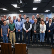 Grupo de aproximadamente 25 pessoas posa para foto em auditório, dispostas em três fileiras. A maioria veste roupas formais, e ao fundo há uma parede com imagem em tons de cinza.