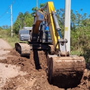 A imagem mostra uma escavadeira hidráulica em operação à beira de uma estrada de chão batido, durante o dia, sob céu azul e ensolarado. A máquina, com braço e caçamba na cor amarela, está escavando uma vala ao lado da via, próxima a um poste de energia elétrica. Há terra removida acumulada ao redor da escavação. O operador, visível dentro da cabine, utiliza a máquina pesada para movimentar o solo. Ao fundo, há vegetação densa e alguns postes de energia alinhados ao longo da estrada.