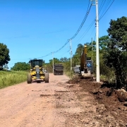 A imagem mostra uma estrada de terra sob céu limpo e azul, onde ocorre uma obra de infraestrutura. À direita, uma escavadeira está operando próxima a um poste de energia elétrica, removendo terra e vegetação da margem da estrada. Um trabalhador pode ser visto dentro da cabine da escavadeira. Há também um caminhão basculante mais ao fundo, aparentemente esperando para carregar ou descarregar materiais. À esquerda da estrada, um motoniveladora (patrol) amarela e preta avança no sentido da câmera, possivelmente nivelando o solo. A via está parcialmente coberta por terra solta, sugerindo que os trabalhos estão em andamento para melhorar ou alargar a estrada. Vegetação verde margeia ambos os lados da via.