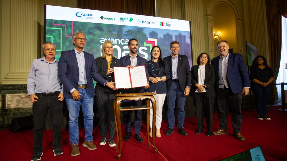 A fotografia mostra um grupo de sete pessoas em pé, posicionadas diante de um painel com a frase “Avançar Mais Cidades”. No centro, está o governador Eduardo Leite segurando um documento aberto, exibido para a câmera.