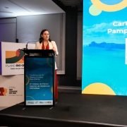 Mulher fala ao microfone em um púlpito sobre um palco, com fundo iluminado por um grande painel digital azul. O painel exibe o texto “Carta do Bioma Pampa (Completa)” e um código QR. Ao lado do púlpito há um banner do “Plano Rio Grande” com o logotipo do Governo do Estado do Rio Grande do Sul.