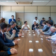 Reunião com autoridades e técnicos em uma sala de reuniões. O governador Eduardo Leite está ao centro, vestindo camisa branca, gesticulando enquanto conversa com o grupo. À sua direita, outro participante fala enquanto os demais ouvem atentos.