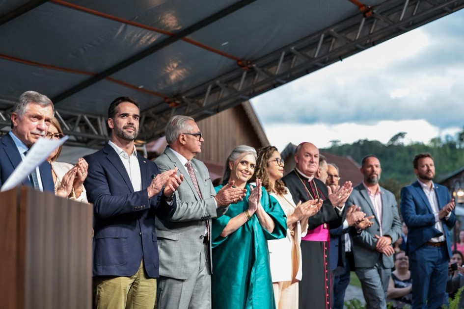 A cena mostra um grupo de pessoas alinhadas sobre um palco. Todos estão de pé, voltados para a frente. As mãos estão posicionadas em gesto de aplauso. Ao fundo, é possível ver parte de uma construção de madeira e uma paisagem com árvores e céu parcialmente nublado.