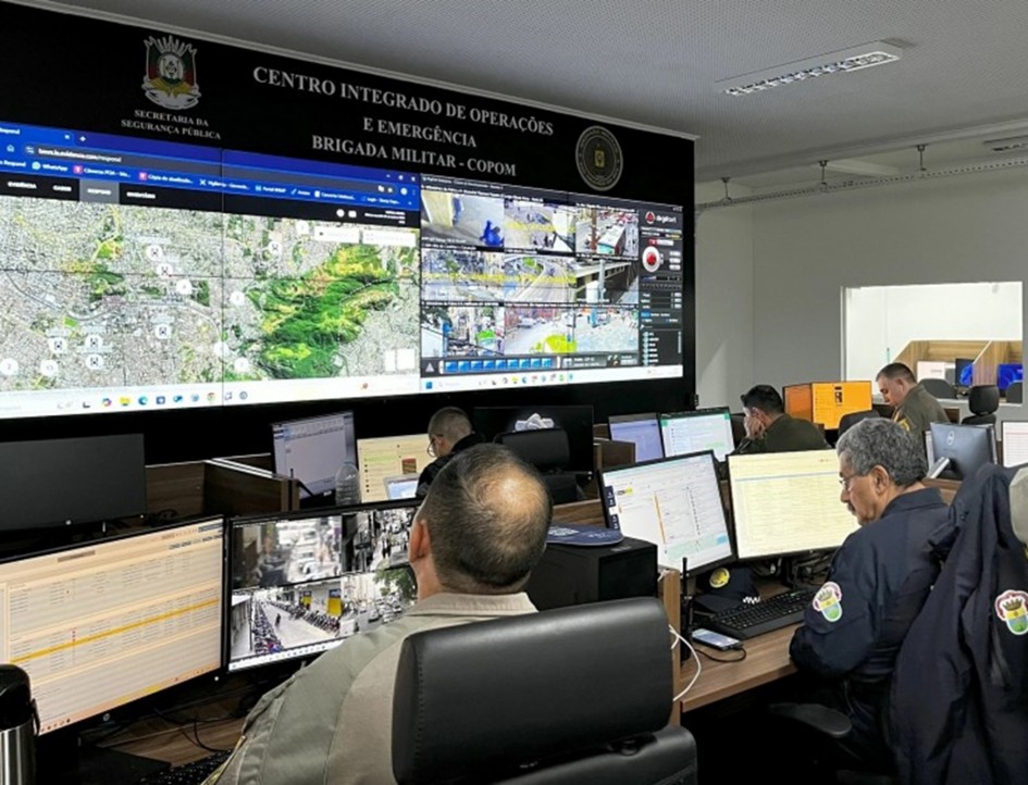 Foto mostra sala de monitoramento do Centro Integrado de Operações. Ao fundo, há um telão com imagem de um mapa e de várias câmeras de monitoramento da cidade. Na sala tem vários agentes trabalhando em seus computadores. Num deles, ele também olha as câmeras.