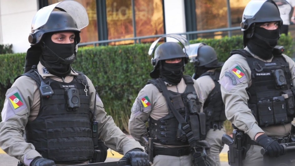 Três policiais usam câmeras corporais. Eles usam máscaras e capacetes.