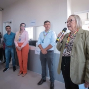 A imagem mostra um grupo de pessoas em um ambiente interno, provavelmente durante a inauguração de uma unidade de saúde pública. Em primeiro plano, à direita, uma mulher idosa de cabelos grisalhos usa óculos, um blazer verde claro e segura um microfone, aparentando estar discursando. Ao fundo, outras pessoas, homens e mulheres, estão em pé, ouvindo atentamente. Elas usam crachás, indicando que podem ser funcionários ou autoridades. Atrás deles, há um balcão com a placa "Recepção" e outra sinalização escrita "Atendimento Geral". O ambiente é claro e limpo, com paredes brancas e piso claro, sugerindo uma instalação recém-inaugurada ou reformada.