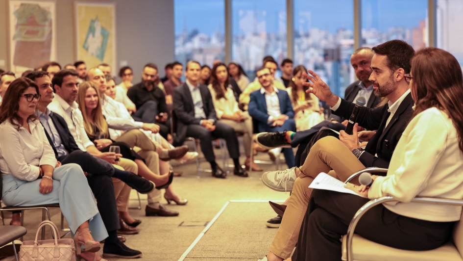 Pessoas auxiliam no painel de discussão em sala moderna com janelas amplas e vista urbana. Palestrantes debatem ativamente, enquanto o público acompanha com atenção. Ambiente profissional e organizado.