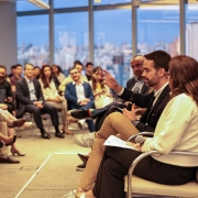 Pessoas auxiliam no painel de discussão em sala moderna com janelas amplas e vista urbana. Palestrantes debatem ativamente, enquanto o público acompanha com atenção. Ambiente profissional e organizado.