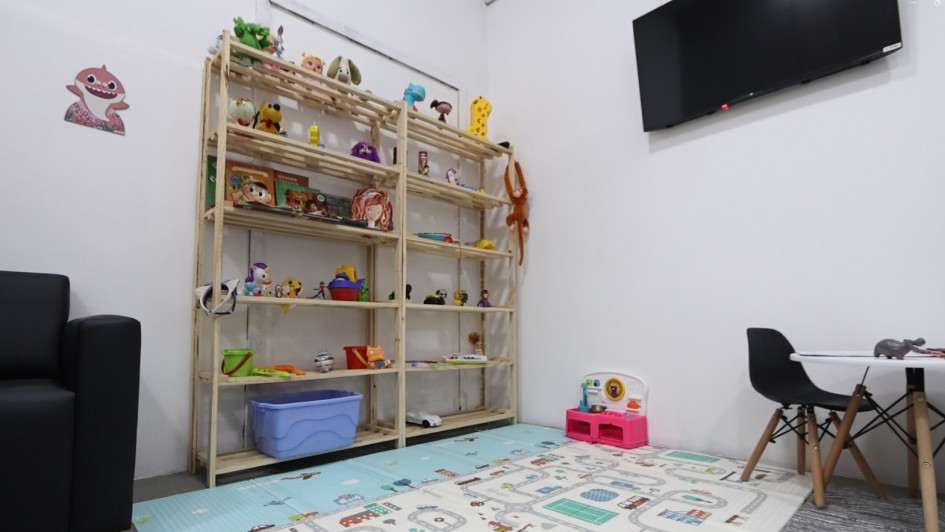 Sala infantil com ambiente acolhedor e brinquedos. À esquerda, há parte de um sofá preto. No centro, uma estante de madeira exibe diversos brinquedos e livros infantis. No chão, um tapete colorido com desenhos de ruas e pistas cobre parte do espaço. À direita, há uma pequena mesa branca com duas cadeiras infantis e brinquedos sobre o tampo. Na parede, acima da estante, há uma televisão preta desligada. O ambiente é bem iluminado, com paredes brancas e aparência organizada e lúdica.