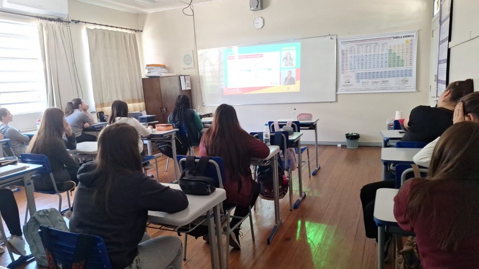 Foto de uma sala de aula, a partir dos fundos do local. Há vários adolescentes, a maioria meninas, sentadas em classes, de costas para a câmera. Elas assistem a aula on-line projetada no quadro branco à frente.
