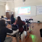 Foto de uma sala de aula, a partir dos fundos do local. Há vários adolescentes, a maioria meninas, sentadas em classes, de costas para a câmera. Elas assistem a aula on-line projetada no quadro branco à frente.