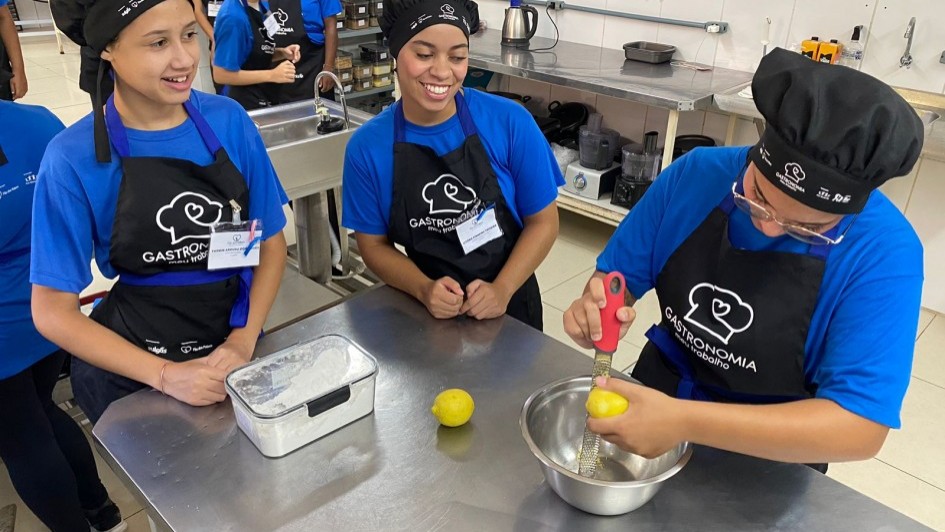 A imagem mostra um grupo de jovens em um ambiente de cozinha profissional, aparentemente participando de uma aula de gastronomia. Três jovens estão em destaque na frente, usando uniformes compostos por camisetas azuis, aventais pretos com o logotipo "Gastronomia" e toucas de chef. Um deles está ralando a casca de um limão sobre uma tigela de metal, enquanto os outros dois observam e sorriem. Em cima da bancada de aço inoxidável há um limão inteiro e um pote transparente com tampa contendo o que parece ser farinha. Ao fundo, outros alunos também uniformizados conversam e observam. Na parede, há uma fileira de facas penduradas, utensílios de cozinha e equipamentos como liquidificadores e chaleiras. O ambiente é limpo e bem iluminado, com um ar profissional e educativo.