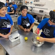 A imagem mostra um grupo de jovens em um ambiente de cozinha profissional, aparentemente participando de uma aula de gastronomia. Três jovens estão em destaque na frente, usando uniformes compostos por camisetas azuis, aventais pretos com o logotipo "Gastronomia" e toucas de chef. Um deles está ralando a casca de um limão sobre uma tigela de metal, enquanto os outros dois observam e sorriem. Em cima da bancada de aço inoxidável há um limão inteiro e um pote transparente com tampa contendo o que parece ser farinha. Ao fundo, outros alunos também uniformizados conversam e observam. Na parede, há uma fileira de facas penduradas, utensílios de cozinha e equipamentos como liquidificadores e chaleiras. O ambiente é limpo e bem iluminado, com um ar profissional e educativo.