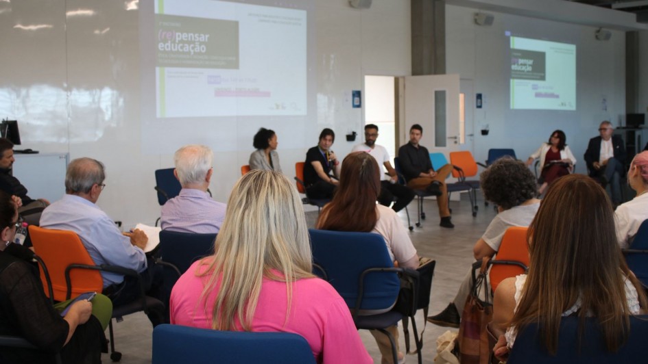 Participantes estão sentados em uma roda de conversa em uma sala moderna e iluminada. No painel projetado, lê-se “(Re)pensar Educação — Ética, tecnologia e humanização na escola”. Educadores e especialistas debatem o tema diante do público atento.