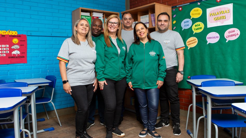 Seis pessoas adultas posam sorridentes dentro de uma sala de aula colorida, com paredes azuis e vermelhas. Três vestem camisetas cinzas e três usam casacos verdes com o logotipo “Educação – Rede Estadual” e a bandeira do Rio Grande do Sul na manga. Ao fundo, há carteiras escolares azuis, cartazes educativos e uma estante com livros.