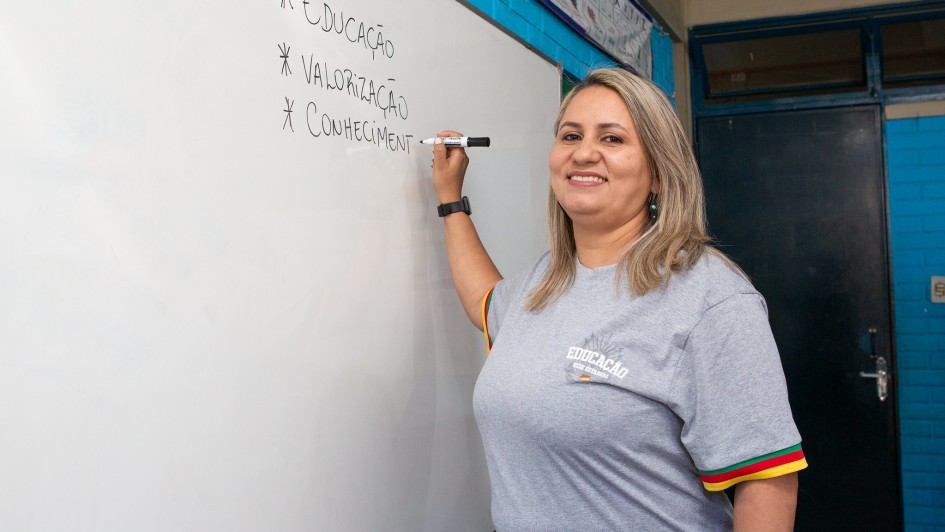 Uma mulher loira de cabelo liso escreve em um quadro branco com caneta preta. Ela usa camiseta cinza com o logotipo “Educação – Rede Estadual” e detalhes com as cores da bandeira gaúcha na manga. No quadro, aparecem as palavras “Educação”, “Valorização” e “Conhecimento”. Ela sorri, olhando para a câmera.