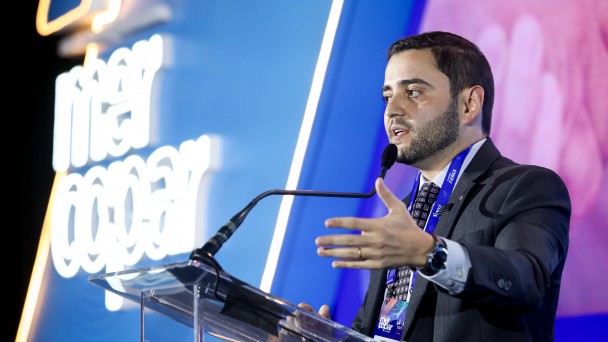 Homem em traje formal fala ao microfone no palco da conferência, com tela iluminada ao fundo exibindo nome do evento. Ambiente profissional e moderno.