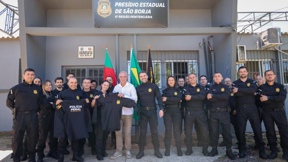 Em frente ao Presídio Estadual de São Borja, um grupo de policiais penais posa para foto oficial com o secretário de Justiça e Sistemas Penal e Socioeducativo, Jorge Pozzobom, de camisa branca. Todos os policiais vestem uniformes pretos e exibem novas camisas da Polícia Penal do Rio Grande do Sul. Ao fundo, bandeiras do Brasil e do Estado completam a cena.