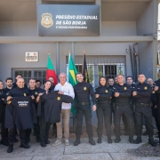 Em frente ao Presídio Estadual de São Borja, um grupo de policiais penais posa para foto oficial com o secretário de Justiça e Sistemas Penal e Socioeducativo, Jorge Pozzobom, de camisa branca. Todos os policiais vestem uniformes pretos e exibem novas camisas da Polícia Penal do Rio Grande do Sul. Ao fundo, bandeiras do Brasil e do Estado completam a cena.