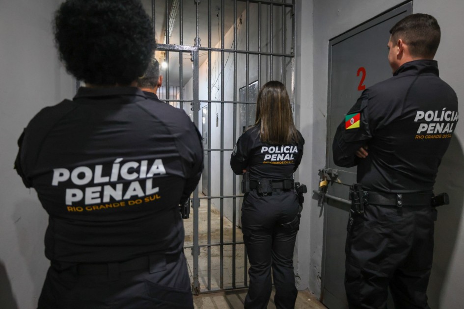 Três policiais penais observam uma cela de segurança no interior do presídio. De costas para a câmera, exibem os novos uniformes com o brasão e o texto “Polícia Penal – Rio Grande do Sul” em destaque nas costas.