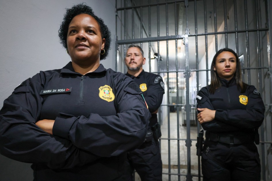 Três policiais penais, dois homens e uma mulher à frente, posam de braços cruzados diante das grades internas de uma galeria prisional. O uniforme preto exibe o emblema dourado da corporação aparece bordado no uniforme.