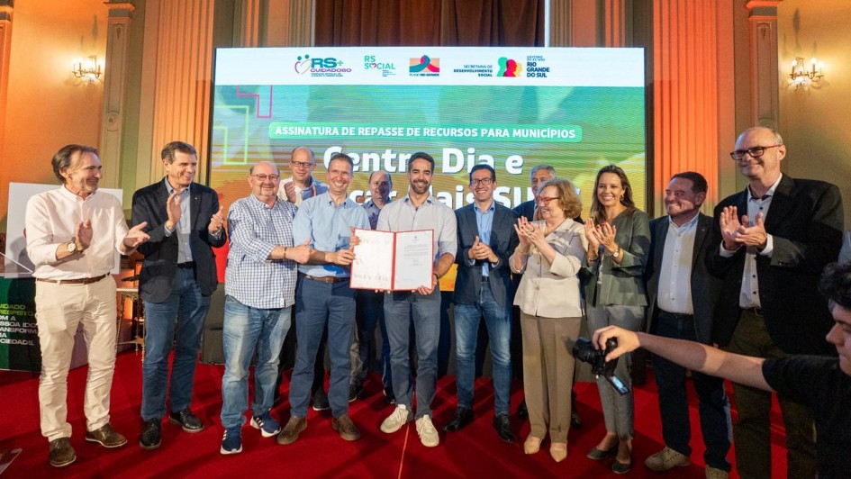 A foto apresenta um grupo de pessoas alinhadas, em frente a um grande painel com a frase “Centro Dia e Avançar Mais SUAS” e logotipos do governo. No centro, duas pessoas seguram um documento aberto. As demais pessoas estão posicionadas lado a lado, algumas com as mãos em gesto de aplauso.
