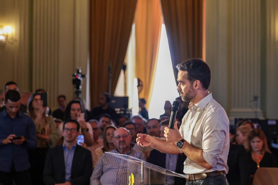 A imagem mostra o governador em pé atrás de um púlpito transparente, segurando um microfone e gesticulando durante a fala. Ao fundo, há várias pessoas sentadas em cadeiras, algumas com celulares, e equipamentos de gravação posicionados.