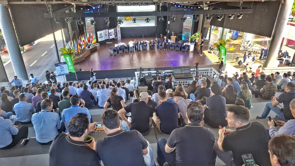 Fotografia de um auditório lotado durante o Milk Summit Brazil, realizado em Ijuí. O público ocupa as arquibancadas, enquanto, no palco, um grupo de palestrantes participa de um painel de debates. Ao fundo, há bandeiras do Brasil e de outros países, além de painéis com logomarcas de patrocinadores e iluminação de palco. A imagem transmite o clima de evento técnico e profissional, com ampla participação de produtores, especialistas e representantes do setor leiteiro.