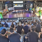 Fotografia de um auditório lotado durante o Milk Summit Brazil, realizado em Ijuí. O público ocupa as arquibancadas, enquanto, no palco, um grupo de palestrantes participa de um painel de debates. Ao fundo, há bandeiras do Brasil e de outros países, além de painéis com logomarcas de patrocinadores e iluminação de palco. A imagem transmite o clima de evento técnico e profissional, com ampla participação de produtores, especialistas e representantes do setor leiteiro.