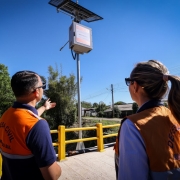 Dois agentes da Defesa Civil, vestindo coletes alaranjados com a inscrição “Defesa Civil – Rio Grande do Sul”, observam e apontam para uma estação de monitoramento recém-instalada em uma ponte. O equipamento possui placa solar e sensores, integrando o sistema de alerta de cheias e eventos climáticos.